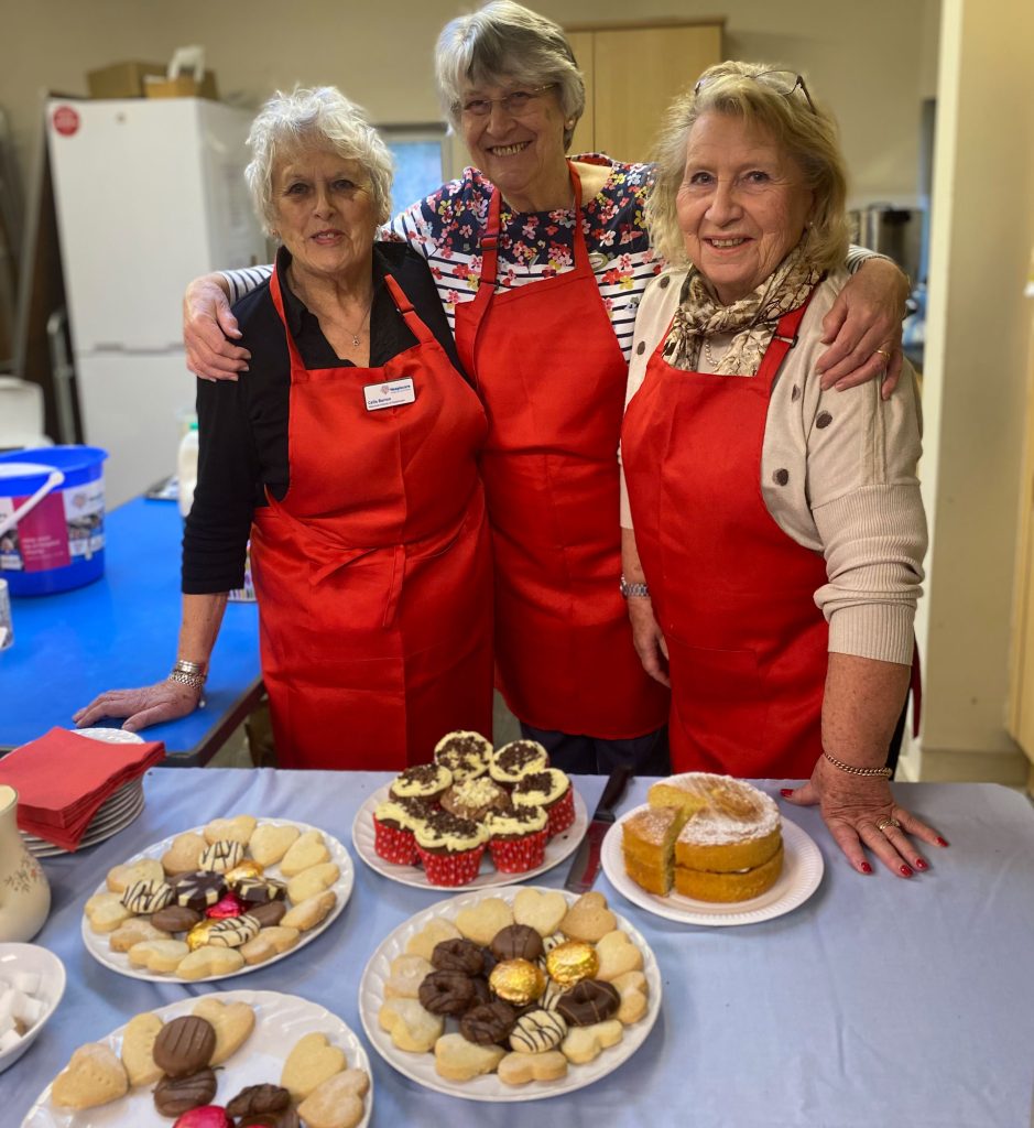 Three women wearing red aprons behind a cake table