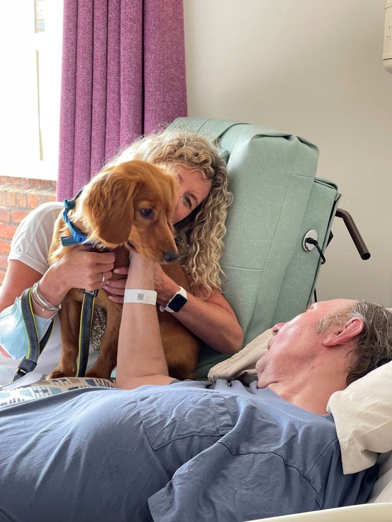 A woman holding a dog and a man in a hospice bed