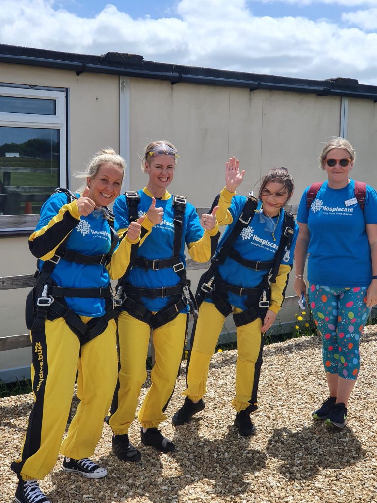 Three women wearing skydive jumpsuits and a fourth woman