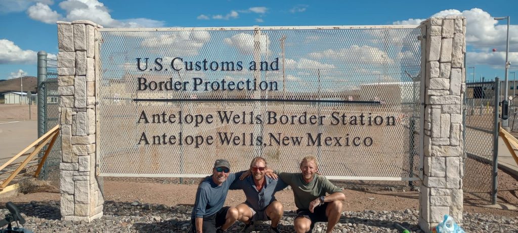 Three men crouched in front of the US/Mexican border sign at Antelope Wells
